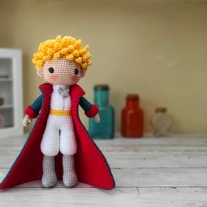 May include: A crocheted doll of The Little Prince, wearing a red cape, a white shirt, and a yellow sash. The doll has blonde hair and a friendly expression.