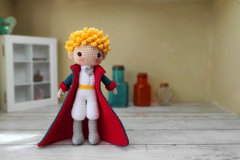 May include: A crocheted doll of The Little Prince, wearing a red cape, a white shirt, and a yellow sash. The doll has blonde hair and a friendly expression.