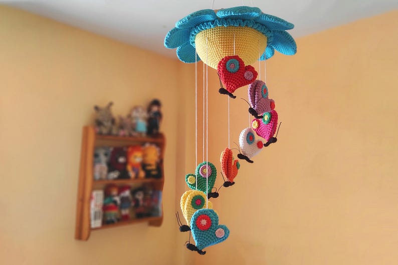 May include: A colourful crocheted baby mobile with a flower-shaped top and hanging butterflies in shades of pink, red, yellow, blue, and green.