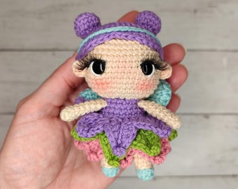 May include: A handmade crochet fairy doll with a light skin tone, large eyelashes, and rosy cheeks. The doll has a purple dress, green and pink skirt, and blue wings. The doll's hair is purple with two small buns on top.