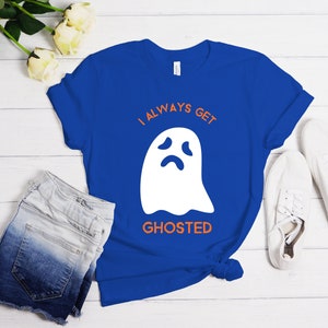 May include: A royal blue t-shirt with the text "I ALWAYS GET GHOSTED" and a white ghost graphic. The shirt is paired with denim shorts and white sneakers. The design is simple and suitable for Halloween or everyday wear.