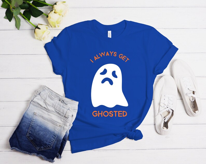 May include: A royal blue t-shirt with the text "I ALWAYS GET GHOSTED" and a white ghost graphic. The shirt is paired with denim shorts and white sneakers. The design is simple and suitable for Halloween or everyday wear.