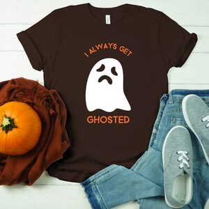 May include: A brown t-shirt with the text "I ALWAYS GET GHOSTED" and a white ghost graphic. The shirt is paired with blue jeans, gray sneakers, and a pumpkin on a brown scarf. A casual Halloween-themed outfit.