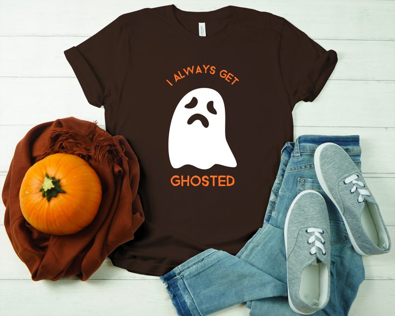May include: A brown t-shirt with the text "I ALWAYS GET GHOSTED" and a white ghost graphic. The shirt is paired with blue jeans, gray sneakers, and a pumpkin on a brown scarf. A casual Halloween-themed outfit.