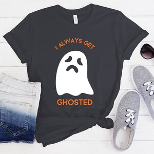May include: Dark gray t-shirt with the text "I ALWAYS GET GHOSTED" in orange above a white ghost graphic. The shirt is paired with denim shorts, gray sneakers, and sunglasses, suggesting a casual, everyday outfit.