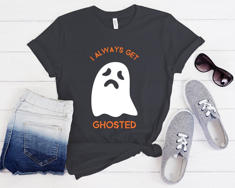 May include: Dark gray t-shirt with the text "I ALWAYS GET GHOSTED" in orange above a white ghost graphic. The shirt is paired with denim shorts, gray sneakers, and sunglasses, suggesting a casual, everyday outfit.