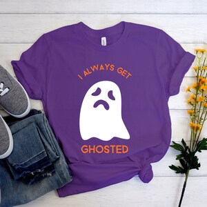 May include: A purple t-shirt with the text "I ALWAYS GET GHOSTED" and a white ghost graphic. The shirt is paired with gray sneakers and blue jeans. Yellow flowers are in the background.