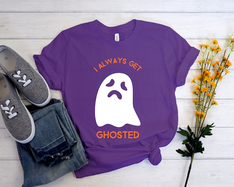 May include: A purple t-shirt with the text "I ALWAYS GET GHOSTED" and a white ghost graphic. The shirt is paired with gray sneakers and blue jeans. Yellow flowers are in the background.