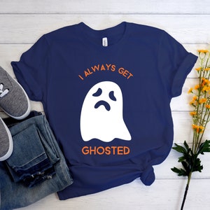 May include: Navy blue t-shirt with the text "I ALWAYS GET GHOSTED" in orange above a white ghost graphic. The shirt is paired with blue jeans and gray sneakers with white laces. Yellow flowers are in the background.