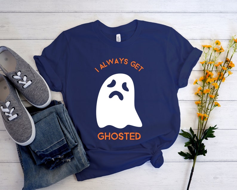 May include: Navy blue t-shirt with the text "I ALWAYS GET GHOSTED" in orange above a white ghost graphic. The shirt is paired with blue jeans and gray sneakers with white laces. Yellow flowers are in the background.