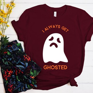 May include: Burgundy t-shirt with the text "I ALWAYS GET GHOSTED" in orange and a white ghost graphic. The shirt is a short-sleeved crew neck style, perfect for Halloween or spooky season.