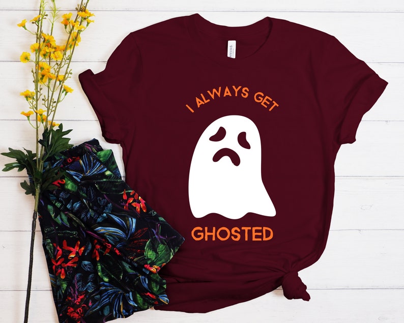 May include: Burgundy t-shirt with the text "I ALWAYS GET GHOSTED" in orange and a white ghost graphic. The shirt is a short-sleeved crew neck style, perfect for Halloween or spooky season.