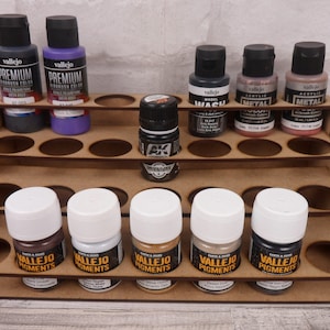 May include: A wooden paint rack with four shelves holds various bottles of acrylic paint, model wash, and pigments. The bottles are arranged in rows and are labelled with the brand name "Vallejo".