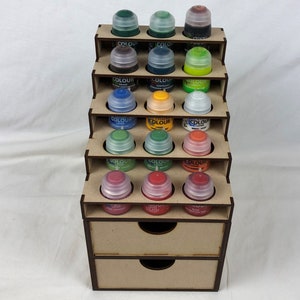 May include: A wooden paint pot storage rack with five shelves and two drawers. The rack is filled with rows of small paint pots with various coloured lids.