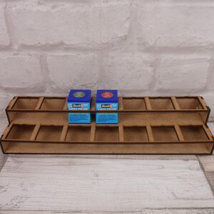 May include: A two-tiered wooden paint rack with 12 slots. The rack is made of light brown wood and has a natural finish. Two small paint bottles are displayed in the top row of the rack. The bottles are blue and green with white labels that read "Revell Aqua Color".