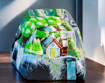 May include: A glass paperweight with a miniature winter scene inside. The scene includes a small house, trees, and a waterfall. The paperweight is clear and has a frosted finish.