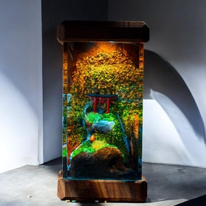 Könnte beinhalten: Eine Holzkiste mit einem transparenten Harzeinsatz, der eine Miniaturlandschaft mit einem kleinen roten Torii-Tor, grünem Laub und einem blauen Wassermerkmal enthält.