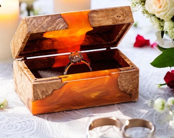 Op de afbeelding: Een houten ringdoosje met een oranje harsinterieur bevat een diamanten verlovingsring. Twee gouden trouwringen liggen op een wit kanten tafelkleed. Brandende kaarsen en een boeket witte rozen en rode rozenblaadjes staan op de achtergrond.