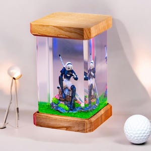 Puede incluir: Una base de lámpara de madera con un cilindro de vidrio transparente que contiene una pequeña figura de una mujer con una espada. La figura está rodeada de hierba verde y flores rosas. La base de la lámpara tiene un acabado marrón claro.