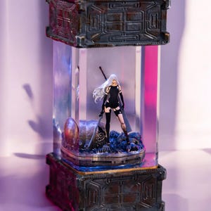 Può includere: Una statuetta di una donna con i capelli bianchi vestita di nero che tiene una spada, in piedi su una base dall'aspetto metallico arrugginito, all'interno di un cubo di vetro trasparente.