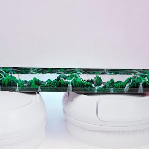 Puede incluir: Una tecla rectangular, verde y blanca, con un diseño de paisaje que presenta árboles y cascadas. La tecla descansa sobre unos auriculares blancos. El diseño de la tecla es verde y blanco.