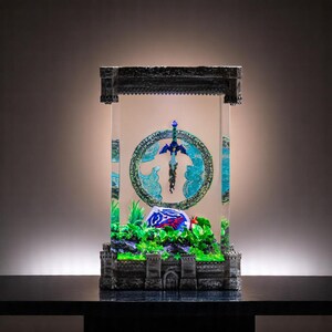 Könnte beinhalten: Eine Vitrine aus Glas mit einem steinartigen Äußeren, die eine Miniatur-Nachbildung des Master Swords aus der Videospielserie The Legend of Zelda zeigt. Das Schwert schwebt in der Luft und ist von einem blauen und grünen Kreisdesign umgeben. Der Sockel der Vitrine ist mit grünen Pflanzen und einer kleinen rot-blauen Figur verziert.
