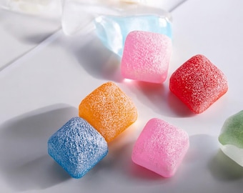 Op de afbeelding: Zes kleurrijke vierkante snoepjes met een korrelige textuur. De snoepjes zijn in tinten blauw, oranje, roze, rood en groen.