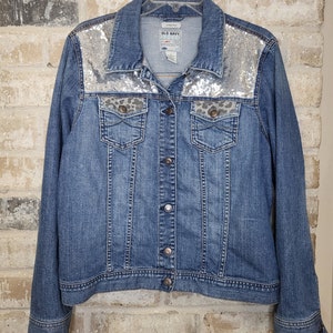 May include: A blue denim jacket with silver sequins on the shoulders and a leopard print design on the pockets. The jacket has a button closure and long sleeves.