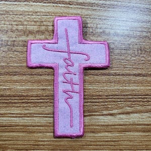May include: A pink cross-shaped patch with the word "faith" embroidered vertically. The patch has a light pink center and a darker pink border. The background is a wooden surface.