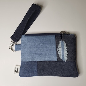 May include: A blue denim clutch purse with a zipper closure and a detachable feather charm. The purse has a black strap and a label that says "Handmade with Love".