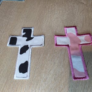 May include: Two cross-shaped fabric patches. The left patch features a white background with black cow-print spots and a silver-toned stitched border. The right patch features a light pink and white camouflage pattern with a dark pink stitched border.