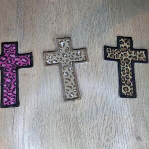 May include: Three cross-shaped patches featuring animal print patterns. The left patch has a pink and black leopard print, the center patch has a light tan and brown leopard print, and the right patch has a classic brown and black leopard print.