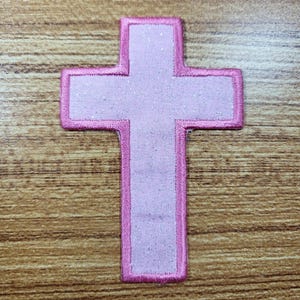 May include: A cross-shaped iron-on patch featuring a light pink fabric center with a subtle glittery finish. The entire perimeter of the cross is outlined with a thick, vibrant pink embroidered border, creating a distinct and clean edge.