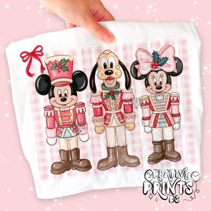 Op de afbeelding: Een wit t-shirt met een grafische print van Mickey Mouse, Pluto en Minnie Mouse verkleed als notenkrakers. De figuren staan tegen een lichtroze ruitjesachtergrond met een rode strik en de tekst Creative Prints DS in de hoek.