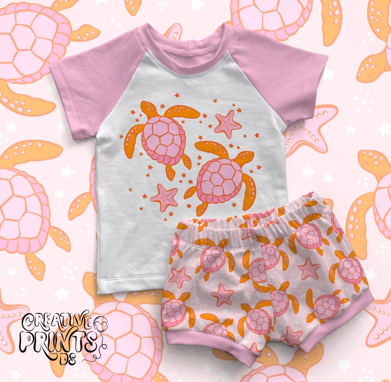 May include: A pink and white two-piece outfit for a child featuring a short-sleeved t-shirt with a graphic of two orange and pink sea turtles and a pair of pink and white shorts with a repeating pattern of orange and pink sea turtles.