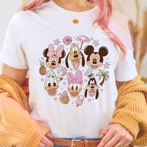 May include: White t-shirt with a colorful graphic print of Mickey Mouse, Minnie Mouse, Donald Duck, Daisy Duck, and Goofy. The characters are surrounded by summer-themed elements like palm trees, pineapples, and beach balls.