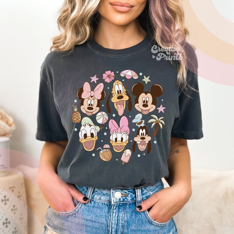 May include: A dark gray t-shirt with a colorful cartoon design featuring Mickey Mouse, Minnie Mouse, Donald Duck, Daisy Duck, and Goofy. The design includes beach-themed elements like palm trees, beach balls, and a pineapple.