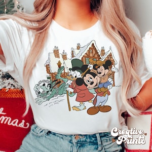May include: A white t-shirt with a vintage-style illustration of Mickey Mouse, Donald Duck, and Goofy in a snowy winter scene. The characters are dressed in winter clothing and are standing in front of a snow-covered house.