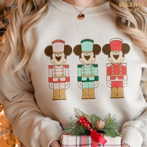 May include: A white sweatshirt with a vintage-style image of three Mickey Mouse characters dressed as nutcrackers. The characters are wearing red, green, and blue uniforms with gold accents.