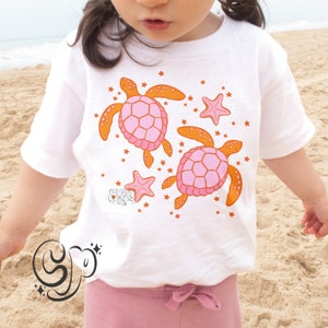 May include: White t-shirt with a pink and orange design of two sea turtles and starfish. The design is on the front of the shirt.