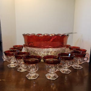 May include: A vintage punch bowl set with a large red glass bowl and matching cups. The bowl has a scalloped rim and sits on a clear glass base. The cups have red rims and handles, perfect for serving beverages at a party.