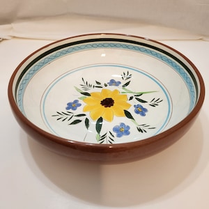 May include: A large, brown ceramic bowl with a white interior and a blue and green floral design. The bowl features a yellow sunflower with blue flowers and green leaves.