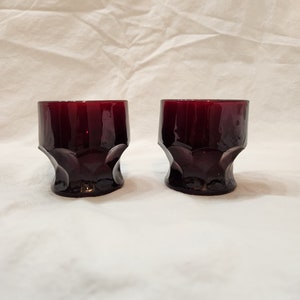 May include: Two dark red glass tumblers with a textured, faceted design. The glasses are empty and sitting on a white surface.