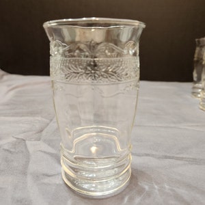 Puede incluir: Un vaso de beber de vidrio transparente con un patr&oacute;n decorativo alrededor de la parte superior e inferior. El vaso tiene una base ancha y una parte superior estrecha.