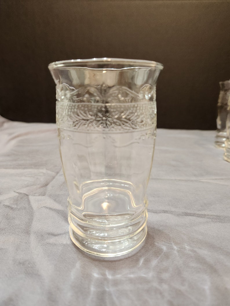 Puede incluir: Un vaso de beber de vidrio transparente con un patr&oacute;n decorativo alrededor de la parte superior e inferior. El vaso tiene una base ancha y una parte superior estrecha.