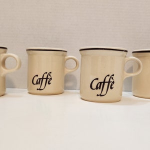 May include: Four white ceramic coffee mugs with black trim and the word "Caffe" written in cursive on the front.