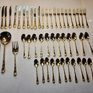 May include: A full set of gold-coloured cutlery, including knives, forks, and spoons, arranged on a white surface. The set includes serving spoons and a serving fork. The handles have a textured floral design.