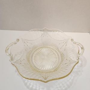 May include: A clear, vintage glass serving dish with a scalloped edge and two handles. The dish features a central, circular design with radiating lines and a decorative leaf pattern. The glass has a slight yellow tint.