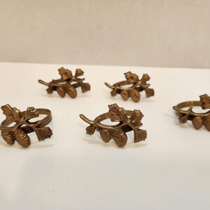 May include: A set of five brass napkin rings, each featuring a detailed metal casting of a pine branch with needles and two small pinecones. The metallic finish has a warm, aged bronze tone with visible texture on the leaves and cones.