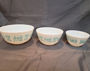 May include: Set of three white mixing bowls with a blue and white folk art design. The bowls are nested together, with the largest bowl in the front and the smallest bowl at the back.